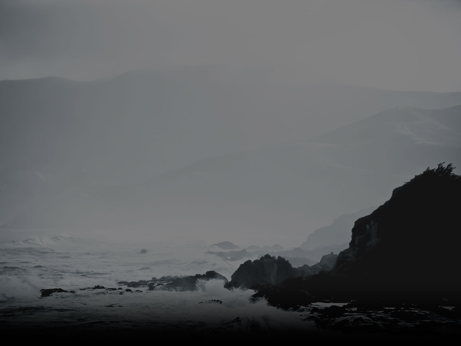 A grayscale coastline at dusk: fog-layered hills behind waves breaking over dark rocks.